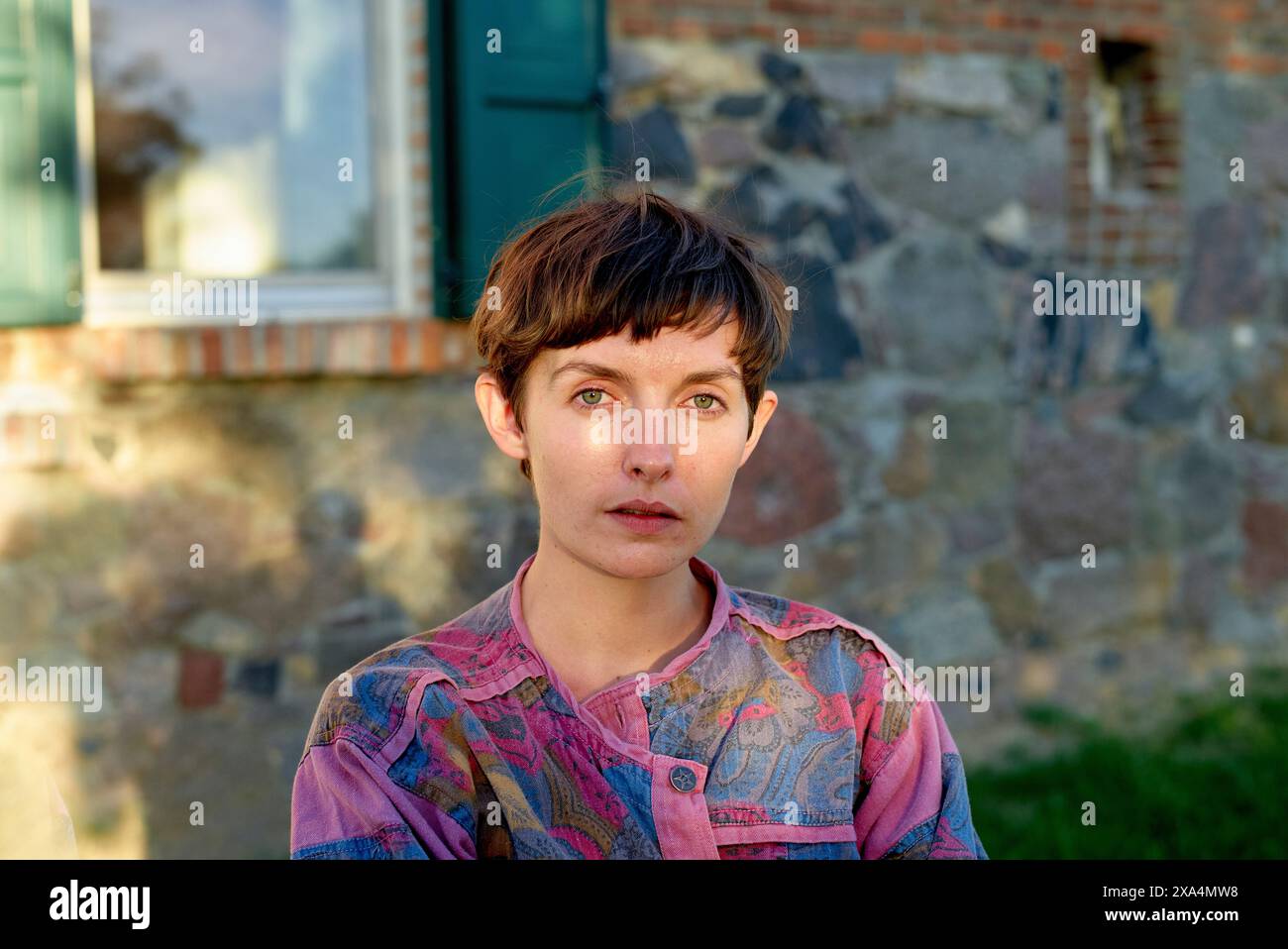 Ein Porträt einer jungen Frau mit kurzen Haaren und auffälligen blauen Augen, die ein gemustertes Hemd trägt, posierte vor einer Steinmauer und einem Fenster. Stockfoto