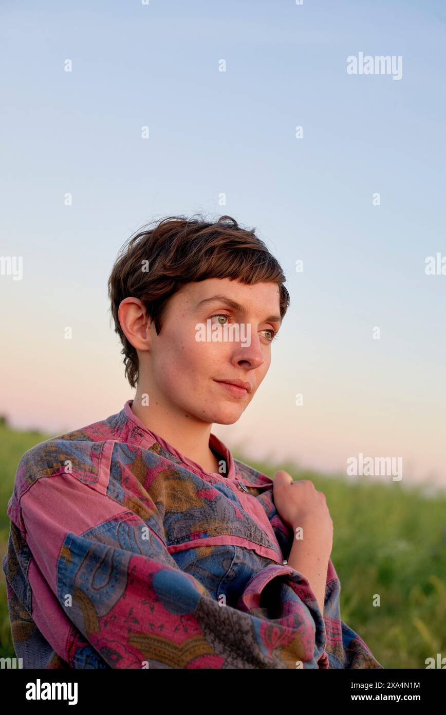 Eine Person mit kurzen Haaren trägt ein bunt bedrucktes Hemd und blickt in der Abenddämmerung mit einem weichen Ausdruck auf einen unscharfen natürlichen Hintergrund. Stockfoto