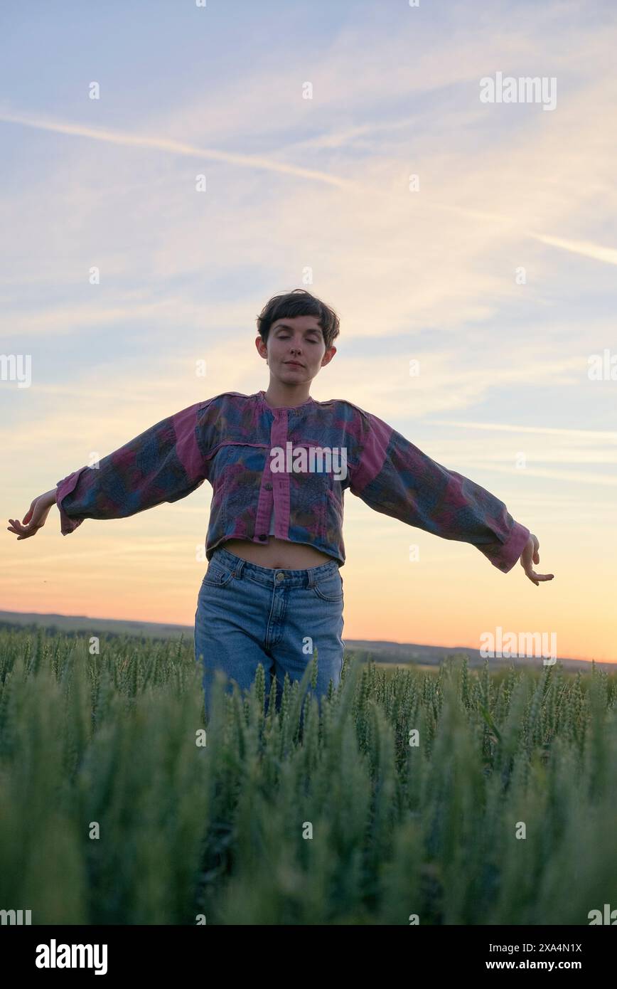 Eine Person steht in der Abenddämmerung in einem hohen Grasfeld mit ausgestreckten Armen und einer entspannten Haltung und trägt eine farbenfrohe Bluse und Jeans. Stockfoto