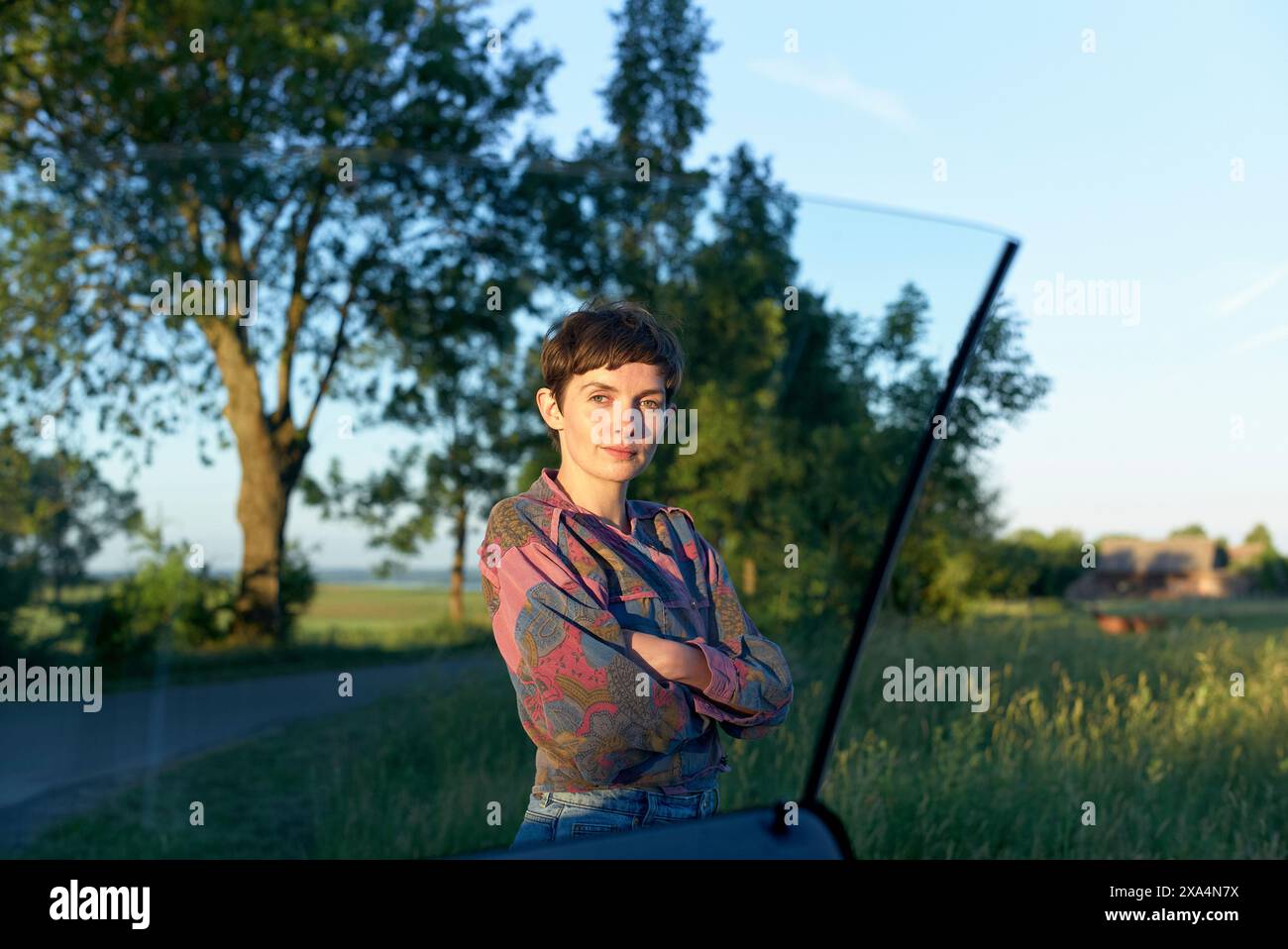 Eine junge Frau steht neben einer offenen Autotür auf einer Landstraße, mit Bäumen und einem Feld im Hintergrund während der goldenen Stunde. Stockfoto