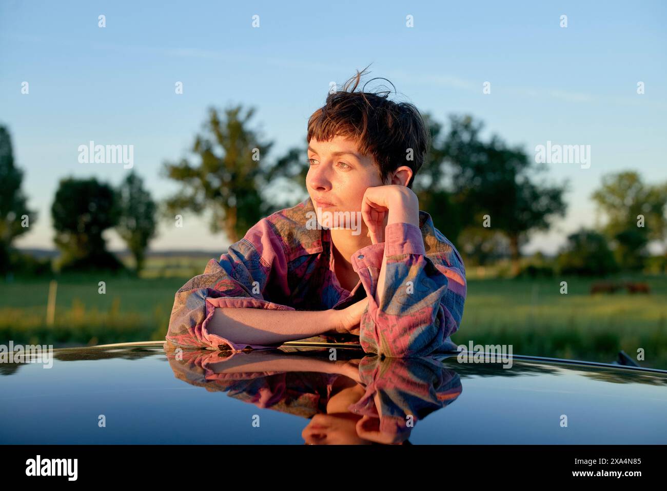 Eine junge Frau mit kurzen Haaren legt ihr Kinn auf die Hand, während sie sich bei Sonnenuntergang auf eine reflektierende Oberfläche lehnt und einen kontemplativen Ausdruck zeigt. Stockfoto