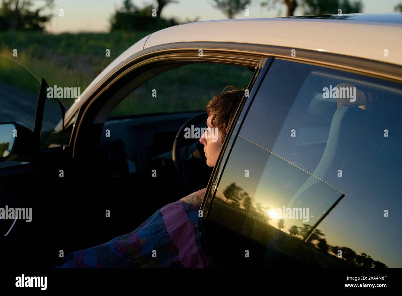 Eine junge Frau sitzt auf dem Fahrersitz eines Autos, deren Silhouette durch das Fenster sichtbar ist, während die Sonne im Hintergrund untergeht und ein warmes Leuchten ausstrahlt. Stockfoto