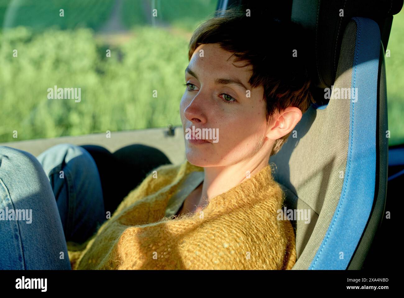 Eine Frau mit kurzen Haaren in einem gelben Pullover sitzt bequem in einem Fahrzeug, mit einem Fenster, das draußen Grün zeigt. Stockfoto