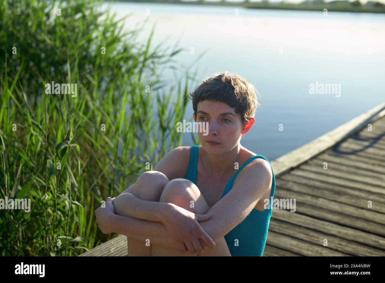 Eine nachdenkliche junge Frau mit kurzen Haaren sitzt auf einem hölzernen Dock an einem Wasserkörper, mit grünem Schilf im Hintergrund und einer ruhigen Seenoberfläche, die das sanfte Licht des Tages reflektiert. Stockfoto