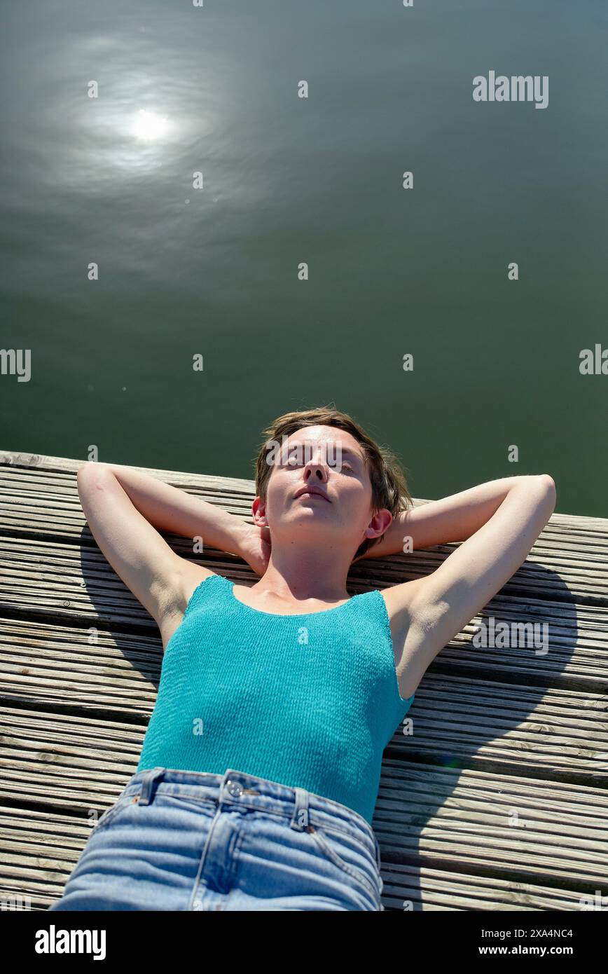 Eine junge Frau liegt auf einer Holzterrasse in der Nähe eines Wassers, sonnt sich im Sonnenlicht mit geschlossenen Augen und Händen hinter dem Kopf. Stockfoto