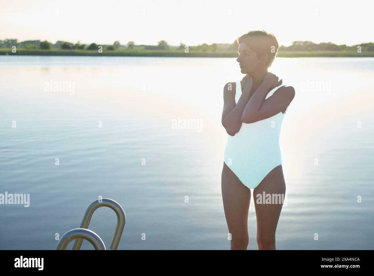Eine Frau steht in der Abenddämmerung am See, trägt einen weißen Badeanzug und sieht nachdenklich aus, während sie mit überkreuzten Armen über das Wasser blickt. Stockfoto