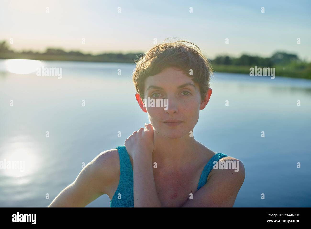 Eine Frau mit kurzen Haaren in einem blauen Kleid steht bei Sonnenuntergang an einem Wasserkörper, wobei das Sonnenlicht ihr Haar sanft hervorhebt. Stockfoto
