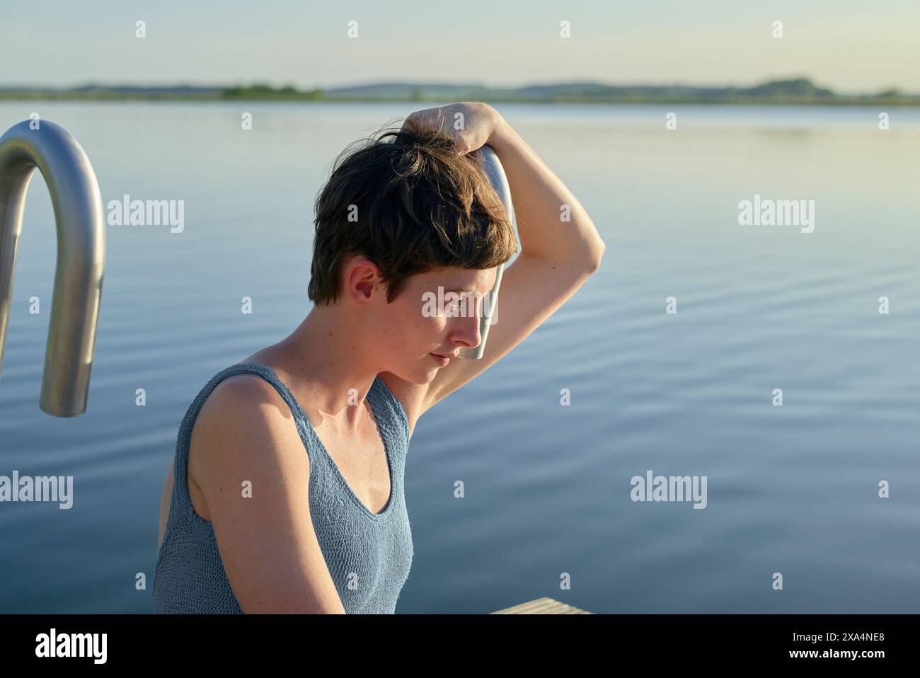 Ein Teenager mit kurzen Haaren legt den Arm an den Kopf und blickt tagsüber nachdenklich über einen ruhigen See. Stockfoto