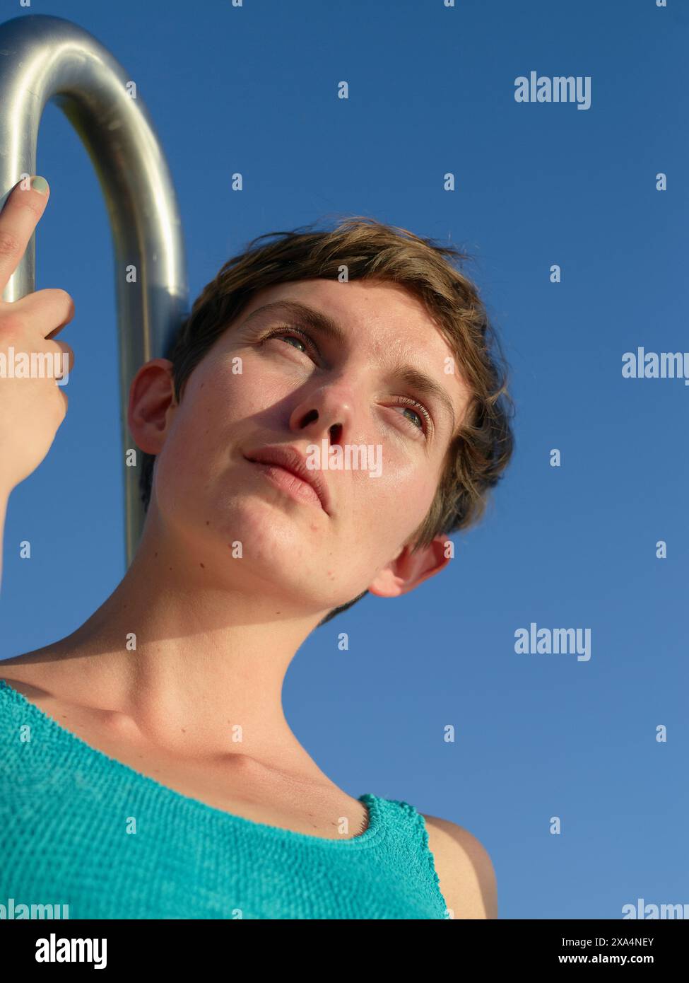 Eine junge Frau mit kurzen Haaren blickt in die Ferne, umrahmt von einem blauen Himmel, mit der Hand auf einer Metallstange. Stockfoto