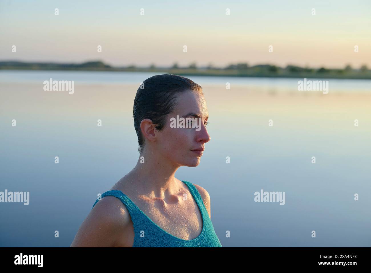 Eine Frau mit nassem Haar, die ein blaublaues Tanktop trägt, blickt in die Ferne an einem ruhigen See in der Abenddämmerung, während warmes Sonnenlicht ihr Profil mit sanften Akzente setzt. Stockfoto