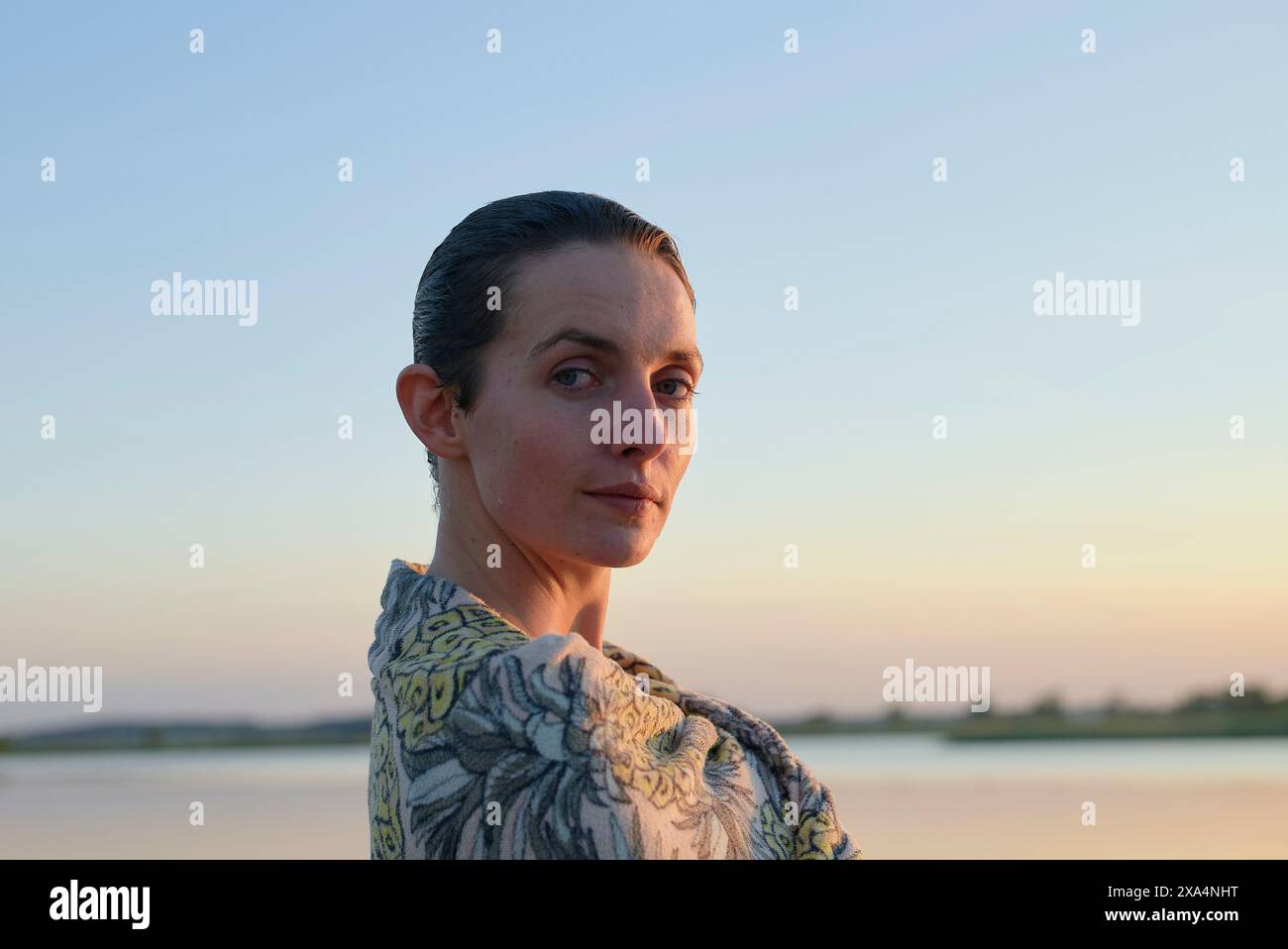 Eine Frau mit nach hinten geglätteten Haaren steht in der Abenddämmerung vor einem ruhigen See und blickt ruhig über ihre Schulter. Stockfoto
