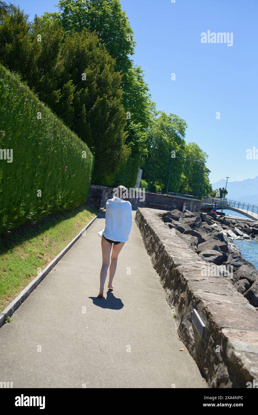 Eine Person spaziert entlang eines gepflasterten Weges an einem See mit üppigem Grün und klarem Himmel im Hintergrund. Stockfoto