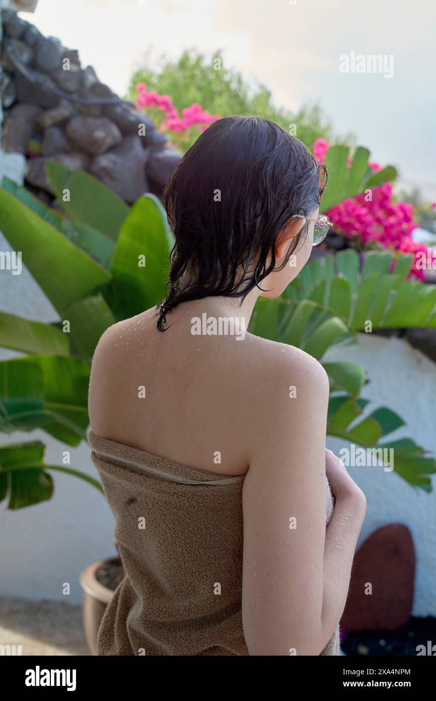 Eine Person wird von hinten gesehen, in ein Handtuch gewickelt, mit nassen Haaren und vor einem Hintergrund von üppigem Grün und leuchtend rosa Blumen. Stockfoto