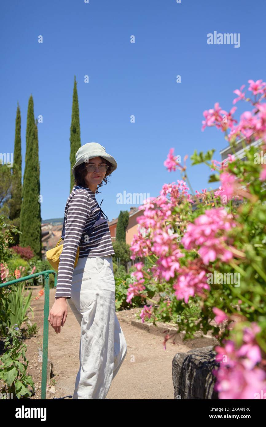 Eine Person in einem gestreiften Oberteil und einer weißen Hose, die auf einem Gartenweg steht, an einem Geländer hält, mit rosa Blumen und Grün im Hintergrund unter einem klaren blauen Himmel. Stockfoto