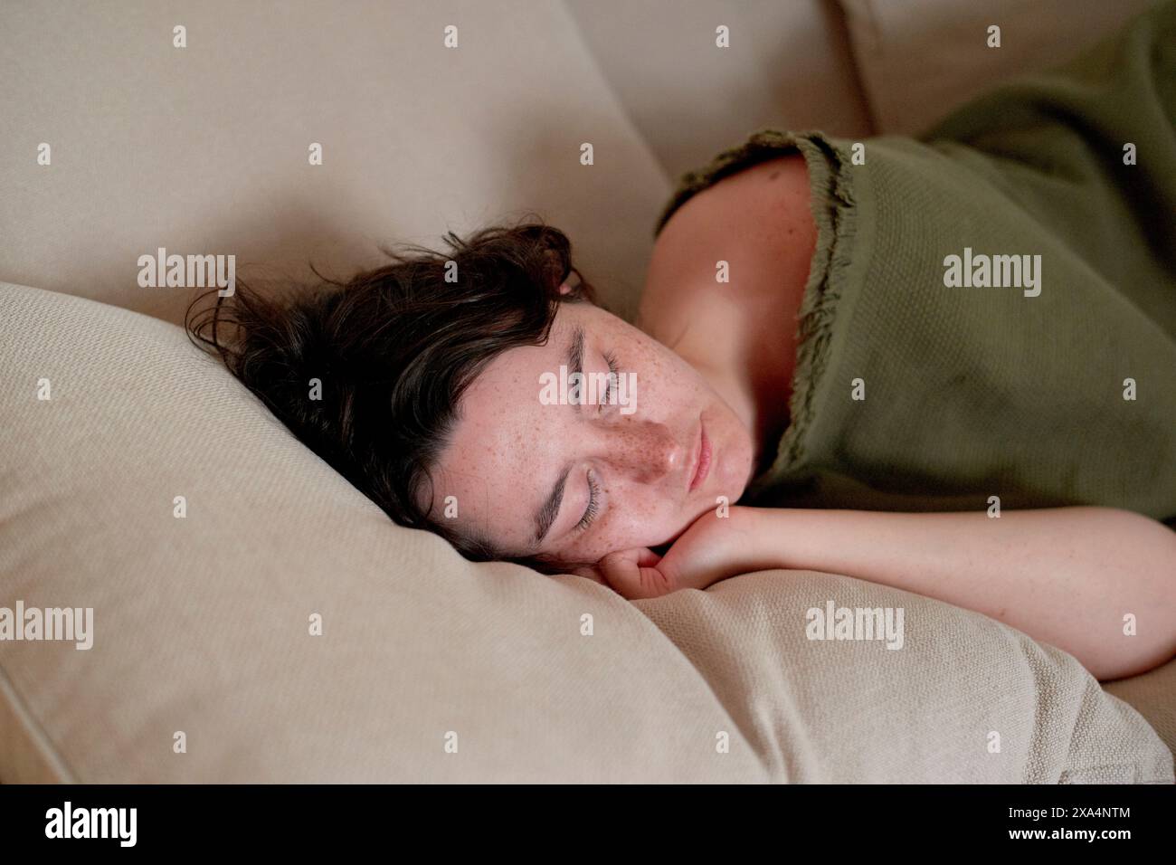 Eine junge Frau schläft friedlich auf einem beigefarbenen Sofa, trägt ein grünes Oberteil, mit dem Kopf auf einem weißen Kissen. Stockfoto