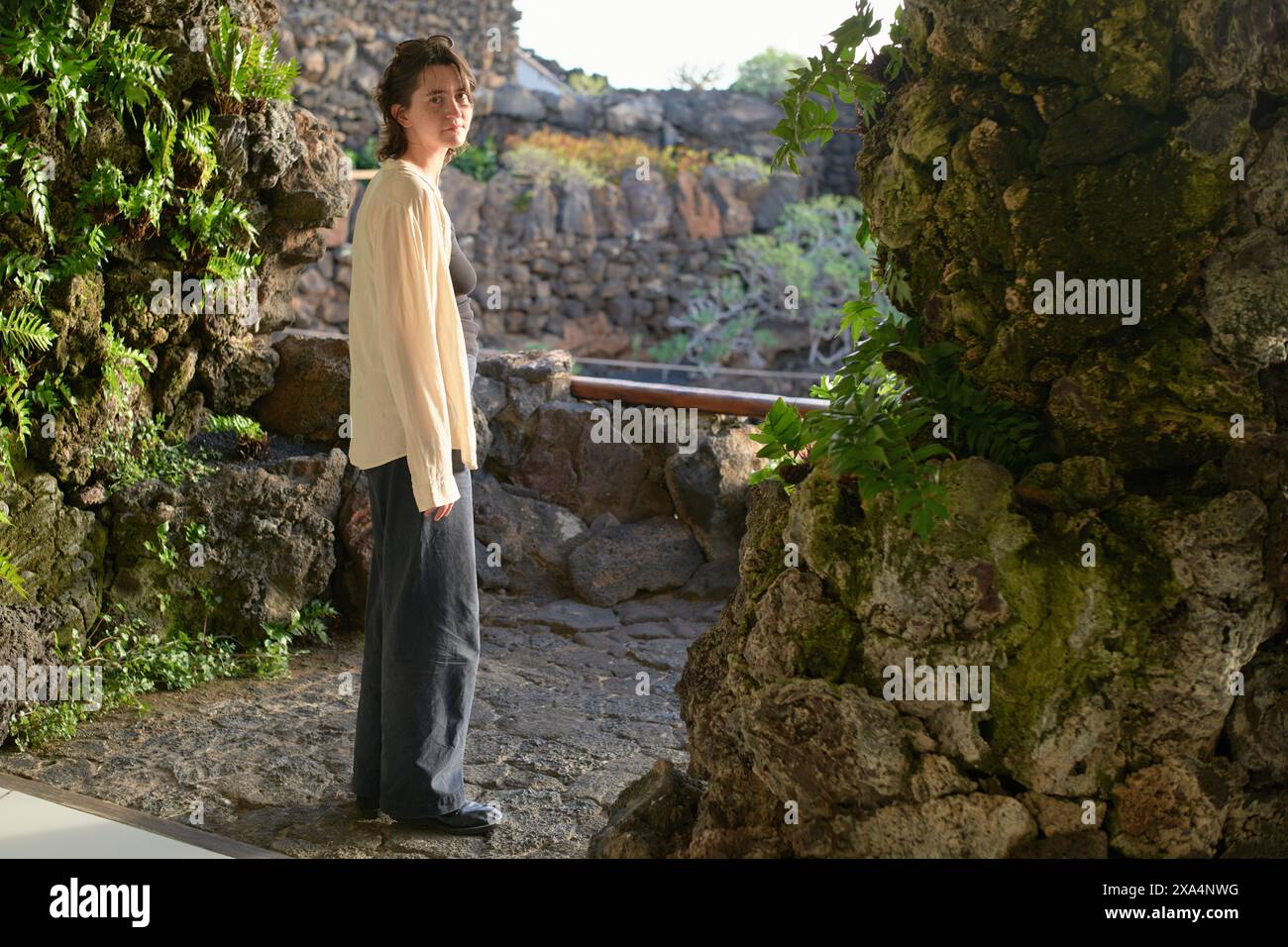 Eine junge Frau steht neben einer Felsmauer und blickt in die Ferne, gekleidet in einem lässigen beigefarbenen Hemd und einer blauen Hose. Stockfoto