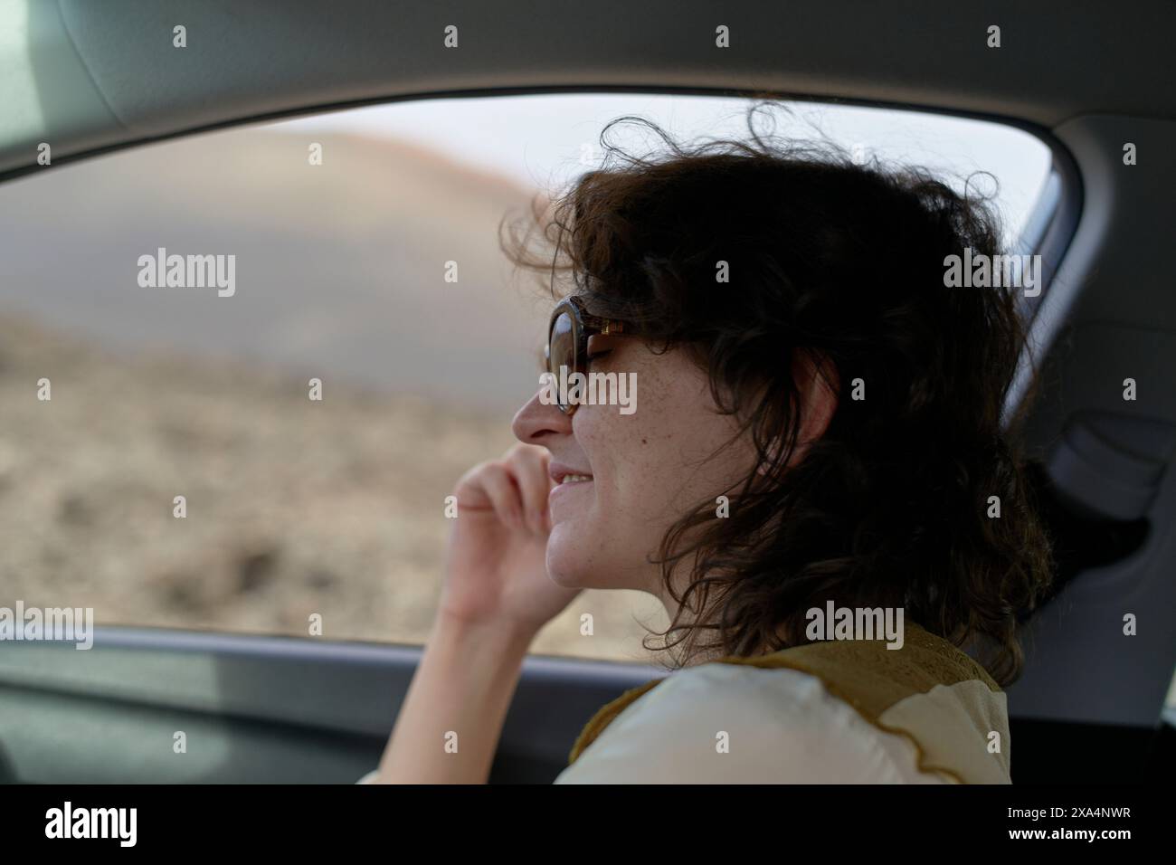 Eine lächelnde Frau mit Sonnenbrille schaut aus dem Fenster eines Autos, wobei ihr Gesicht teilweise von Sonnenlicht beleuchtet wird. Stockfoto