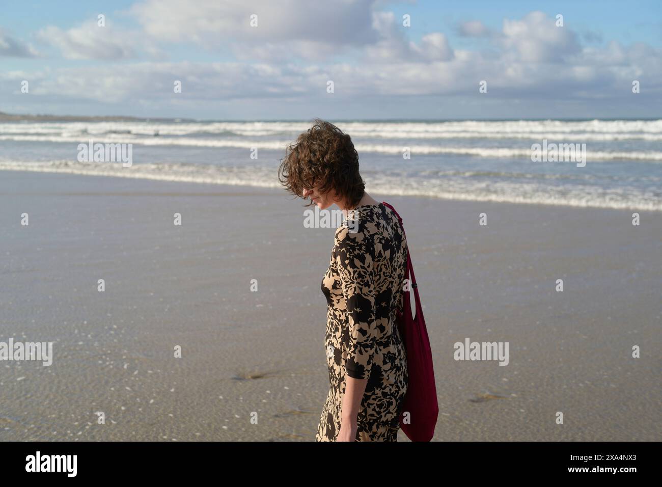 Eine junge Frau mit schulterlangem Haar, trägt ein Kleid und trägt eine rote Tasche, steht an einem Sandstrand mit Wellen im Hintergrund. Stockfoto