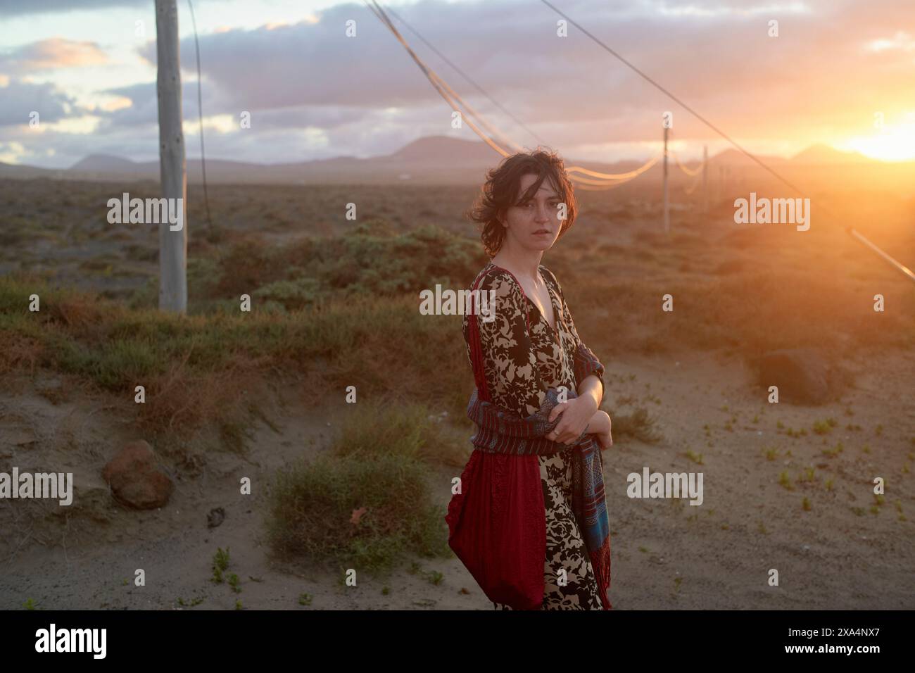 Eine Frau im Bohème-Stil steht bei Sonnenuntergang in einer rauen Landschaft mit Stromleitungen, die sich bis in die Ferne erstrecken. Stockfoto