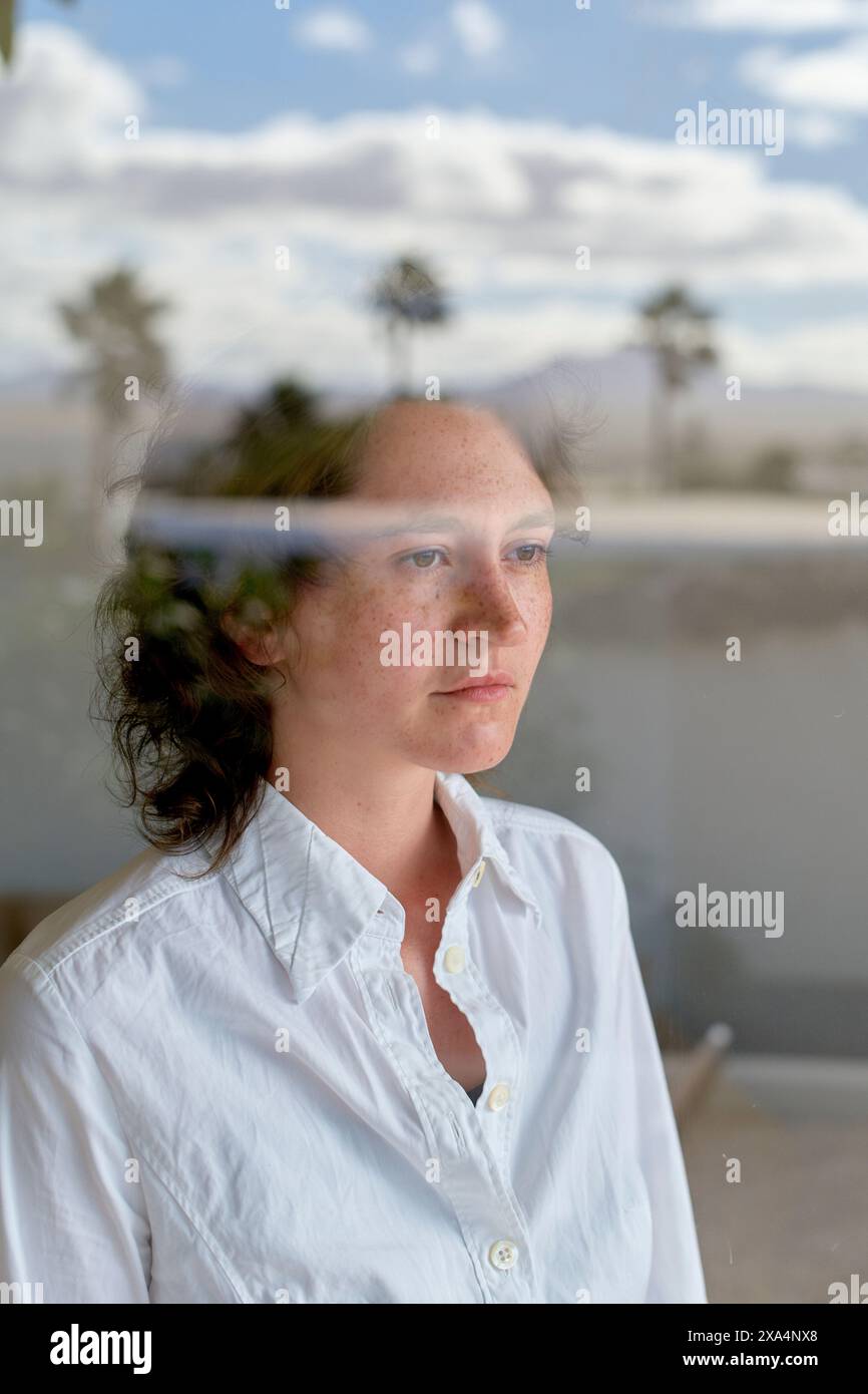 Eine nachdenkliche junge Frau mit lockigem Haar blickt aus einem Fenster, ihre Reflexion und ferne Bäume mischen sich im Bild. Stockfoto
