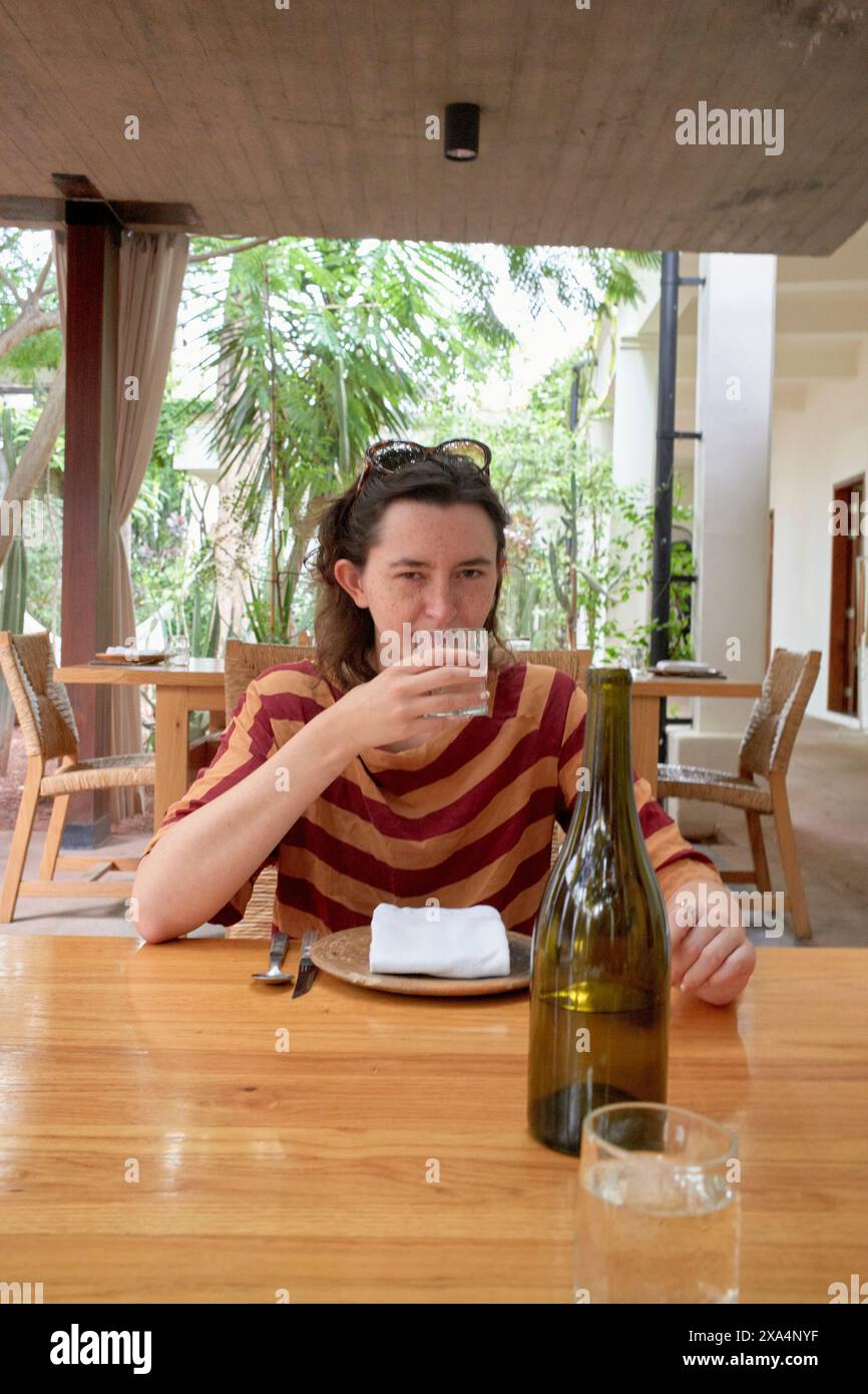 Eine junge Frau, die an einem Esstisch im Freien sitzt, Wasser aus einem klaren Glas trinkt, mit einer leeren Weinflasche im Vordergrund und üppigem Grün im Hintergrund. Stockfoto
