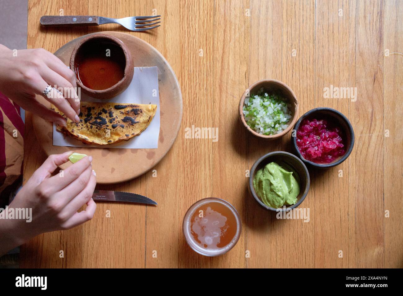 Hände einer Person, die Soße auf einer Quesadilla mit verschiedenen Beilagen und Belägen auf einem Holztisch verteilt. Stockfoto