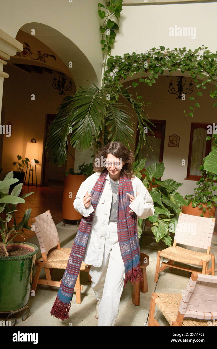 Eine lächelnde junge Frau geht durch einen Innenraum mit Topfpflanzen, hält einen farbenfrohen Schal und trägt legere, stilvolle Kleidung. Stockfoto