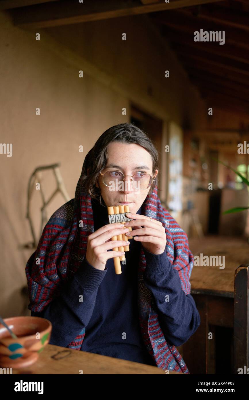 Eine junge Frau sitzt an einem rustikalen Holztisch und spielt eine hölzerne Pfannflöte. Sie tragen eine Brille, einen karierten Schal und haben einen kontemplativen Ausdruck. Stockfoto