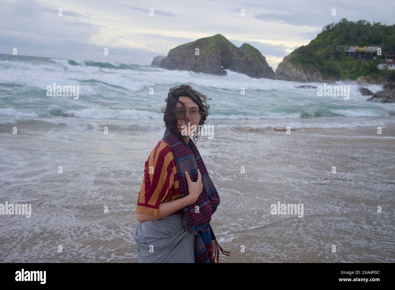 Eine Person steht an einem Sandstrand mit Wellen, die an Land krachen, mit einem Lächeln auf die Kamera gerichtet und trägt ein gestreiftes Oberteil und einen Schal, mit einem felsigen Felsvorsprung und bewölktem Himmel im Hintergrund. Stockfoto