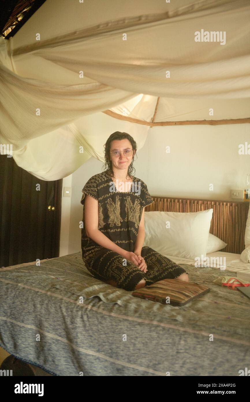 Eine junge Frau sitzt im Kreuz auf einem Bett mit gemusterter Decke in einem mit Moskitonetz bedeckten Zimmer und lächelt bescheiden in die Kamera. Stockfoto