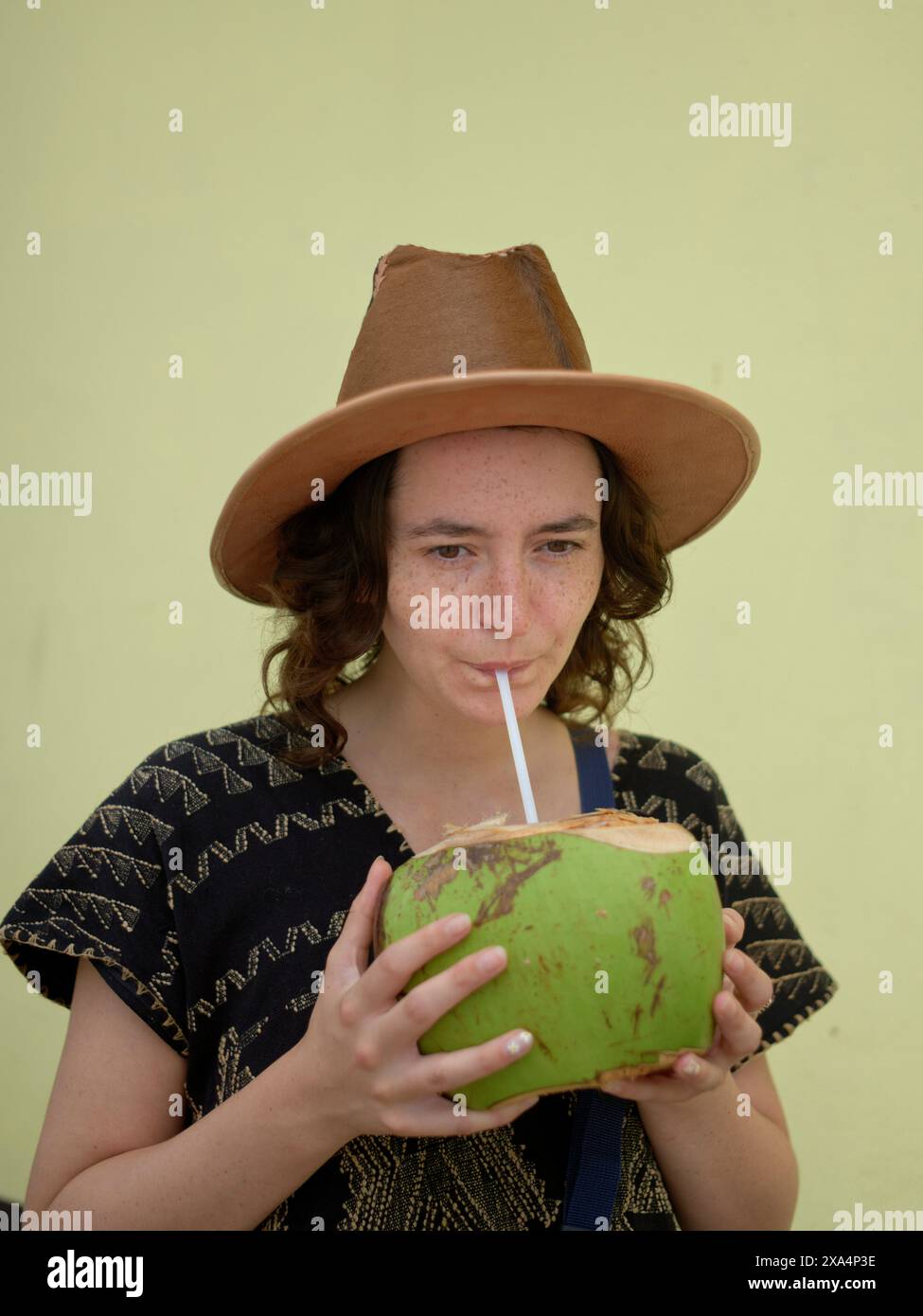 Eine junge Frau, die einen braunen Hut trägt, schlürft an einer Kokosnuss mit einem Strohhalm vor einem blassgelben Hintergrund. Stockfoto