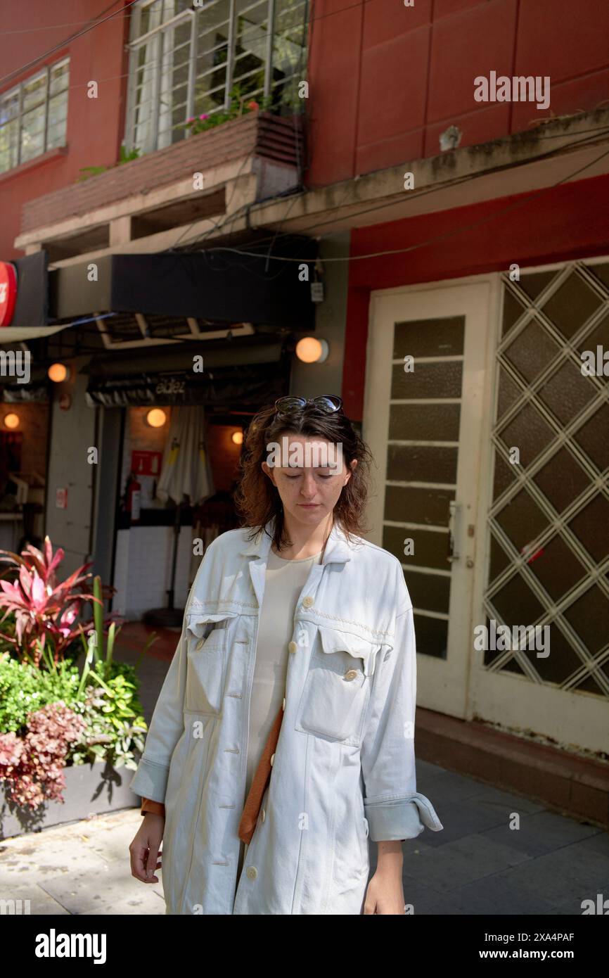 Eine Frau läuft auf einer Stadtstraße vorbei an einem roten Gebäude mit Pflanzen und dem Außenbereich eines Cafés im Hintergrund. Stockfoto