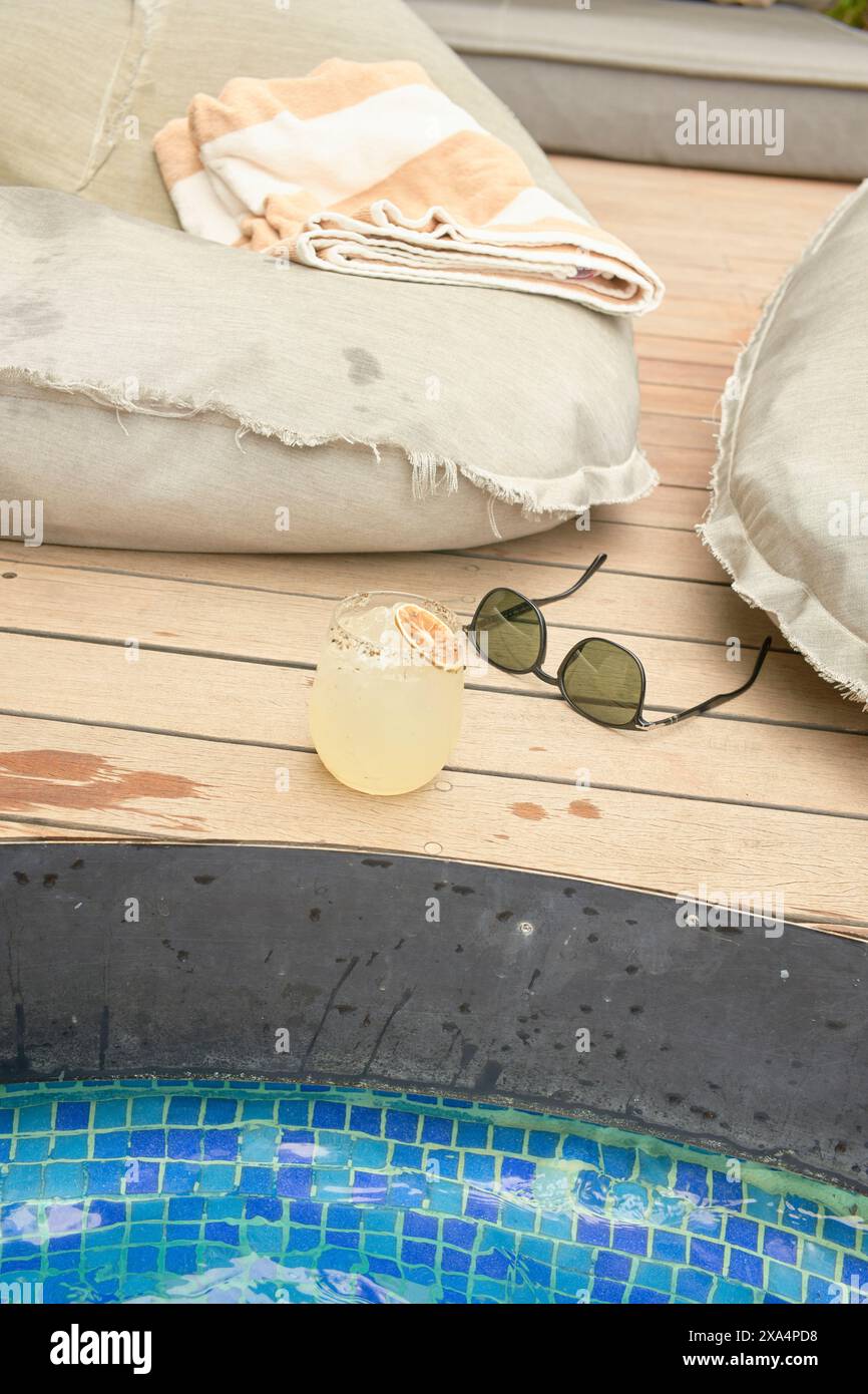 Ein erfrischendes Getränk befindet sich auf einer hölzernen Poolterrasse neben einer Sonnenbrille, mit weichen Sitzgelegenheiten und einem Handtuch im Hintergrund, was eine entspannende Lage am Pool andeutet. Stockfoto