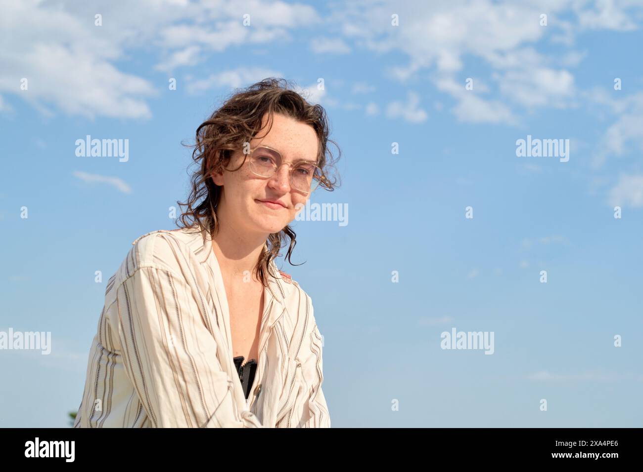 Eine lächelnde Frau mit lockigem Haar, die eine Brille und ein helles gestreiftes Hemd trägt, steht vor einem klaren blauen Himmel. Stockfoto