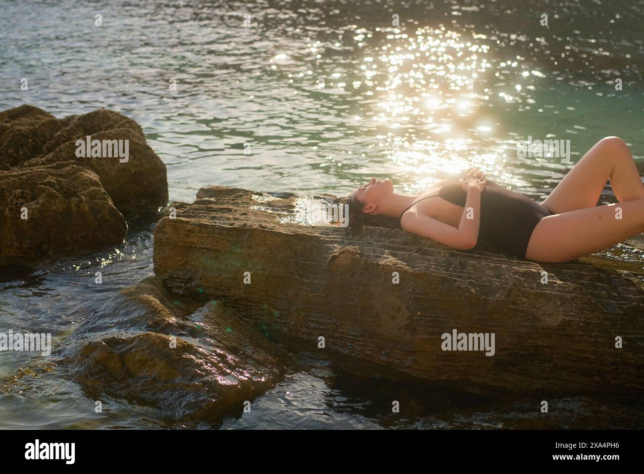 Eine junge Frau liegt auf einem Felsen am Meer und taucht in der Sonne auf, während ihre Hände auf ihrem Mittelteil liegen. Stockfoto
