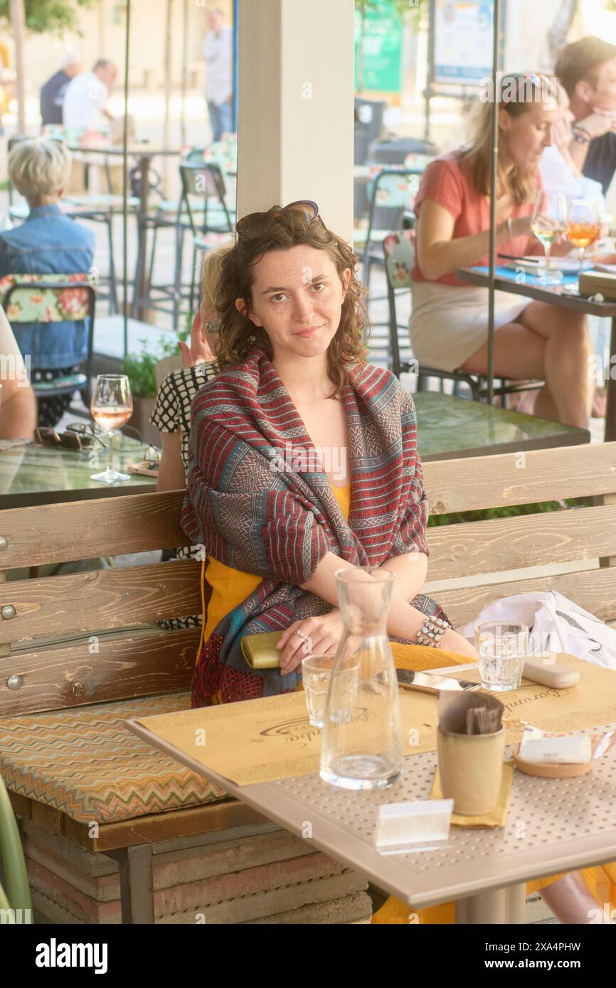 Eine Frau sitzt an einem Holztisch in einem Restaurant im Freien, andere Gäste sind im Hintergrund zu sehen. Stockfoto