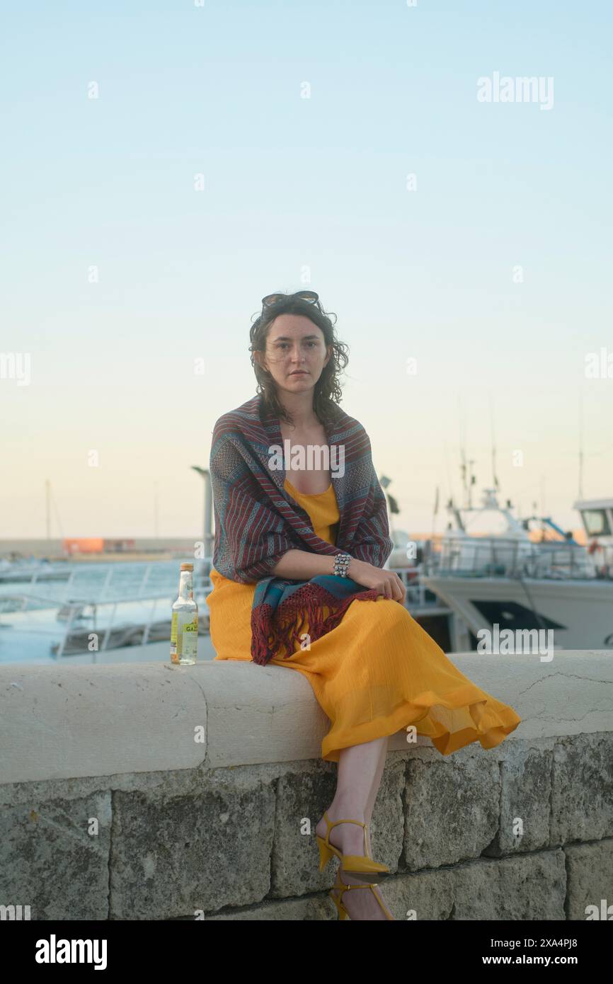 Eine Frau, die in der Abenddämmerung auf einem Steinvorsprung an einem Jachthafen sitzt, ein gelbes Kleid mit einem bunten Schal trägt, während sie einen Drink hält. Stockfoto