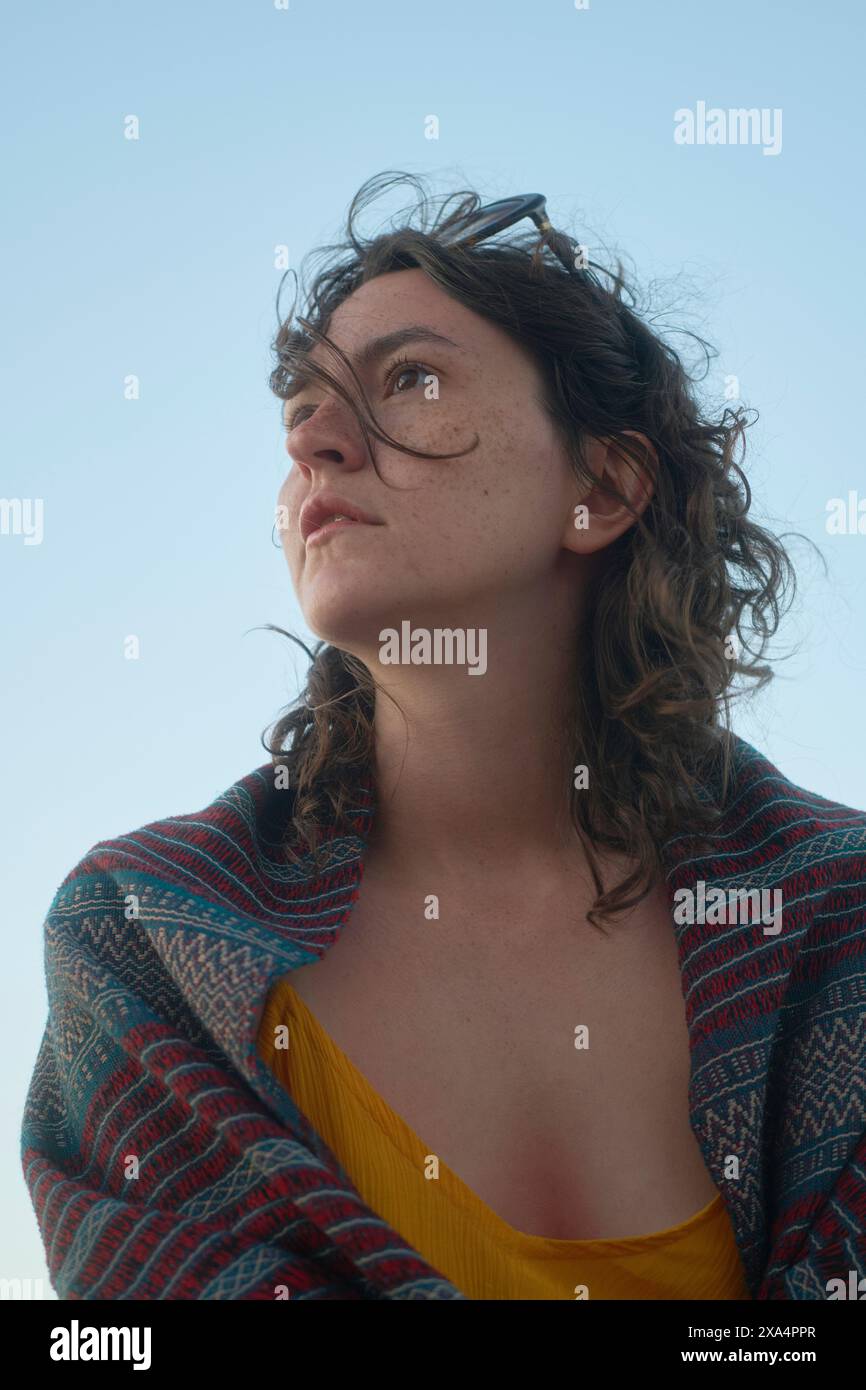 Eine Frau mit lockigem Haar, Sonnenbrille auf dem Kopf und gemusterter Jacke über einem gelben Oberteil, blickt unter klarem Himmel in die Ferne. Stockfoto