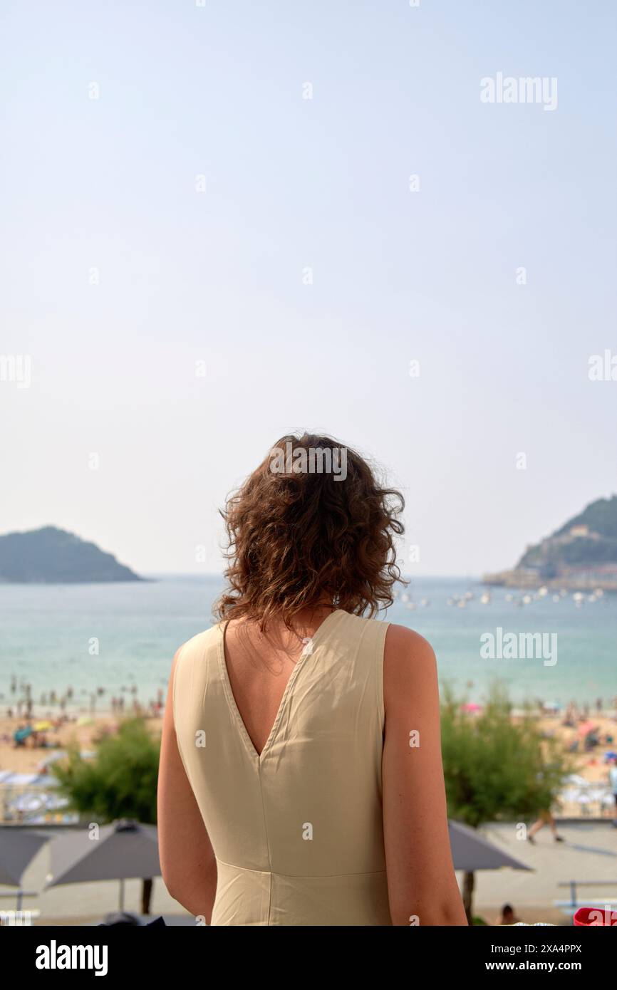 Eine Frau schaut auf eine Strandszene mit klarem Himmel, Sandstränden und fernen Hügeln. Ihr Rücken ist zur Kamera und sie trägt ein ärmelloses Kleid. Stockfoto