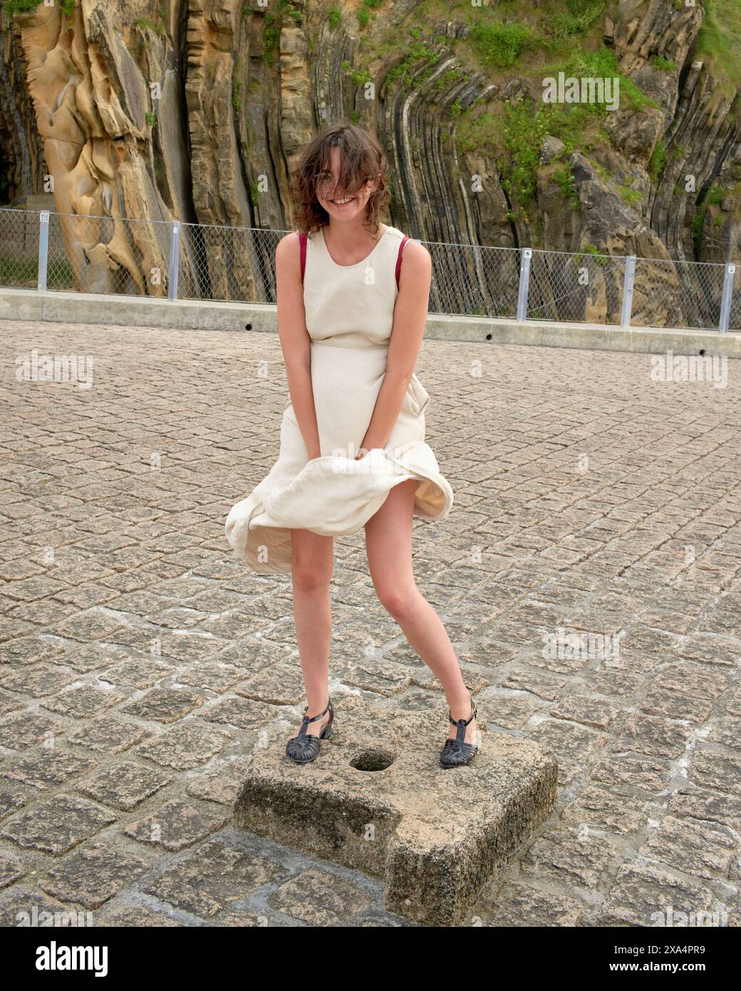 Eine Frau in einem ärmellosen cremefarbenen Kleid steht auf einer Steinplattform mit verspielter Pose, ihr Kleid flattert im Wind, mit einer einzigartigen Felsformation im Hintergrund. Stockfoto