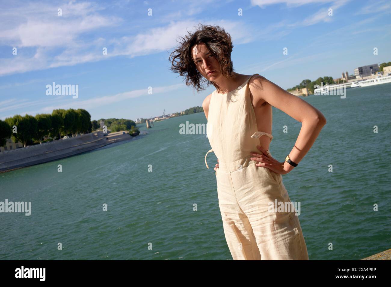Eine Person steht selbstbewusst am Flussufer mit im Wind wehenden Haaren und trägt einen hellen Jumpsuit vor einem blauen Himmel. Stockfoto