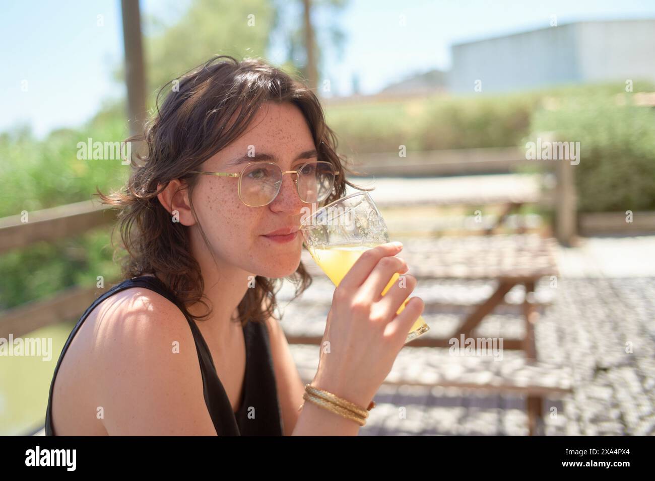 Eine Frau sitzt draußen, schlürft an einem Glas eines blassgelben Getränks, mit Bäumen und einem Gebäude im Hintergrund. Stockfoto