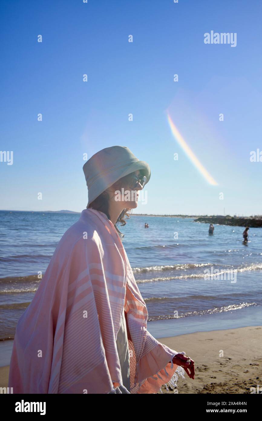 Eine Person schlendert am Strand entlang mit einem hellen Sonnenlicht, das am klaren blauen Himmel zu sehen ist. Stockfoto