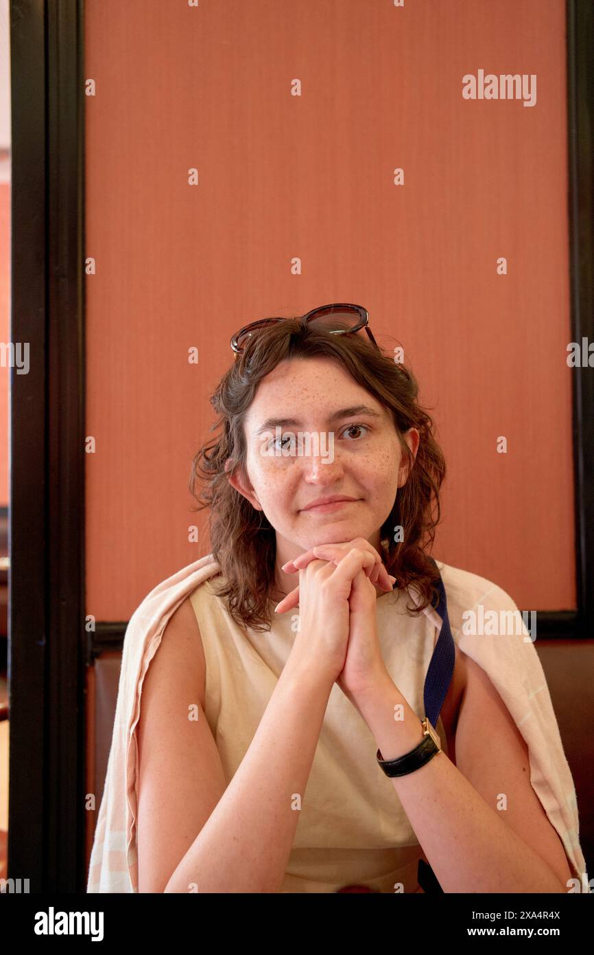 Eine Frau mit Sommersprossen, die ein helles Oberteil und eine Sonnenbrille auf dem Kopf trägt, sitzt drinnen mit dem Kinn auf ihren Händen und blickt mit einem leichten Lächeln in die Kamera. Stockfoto
