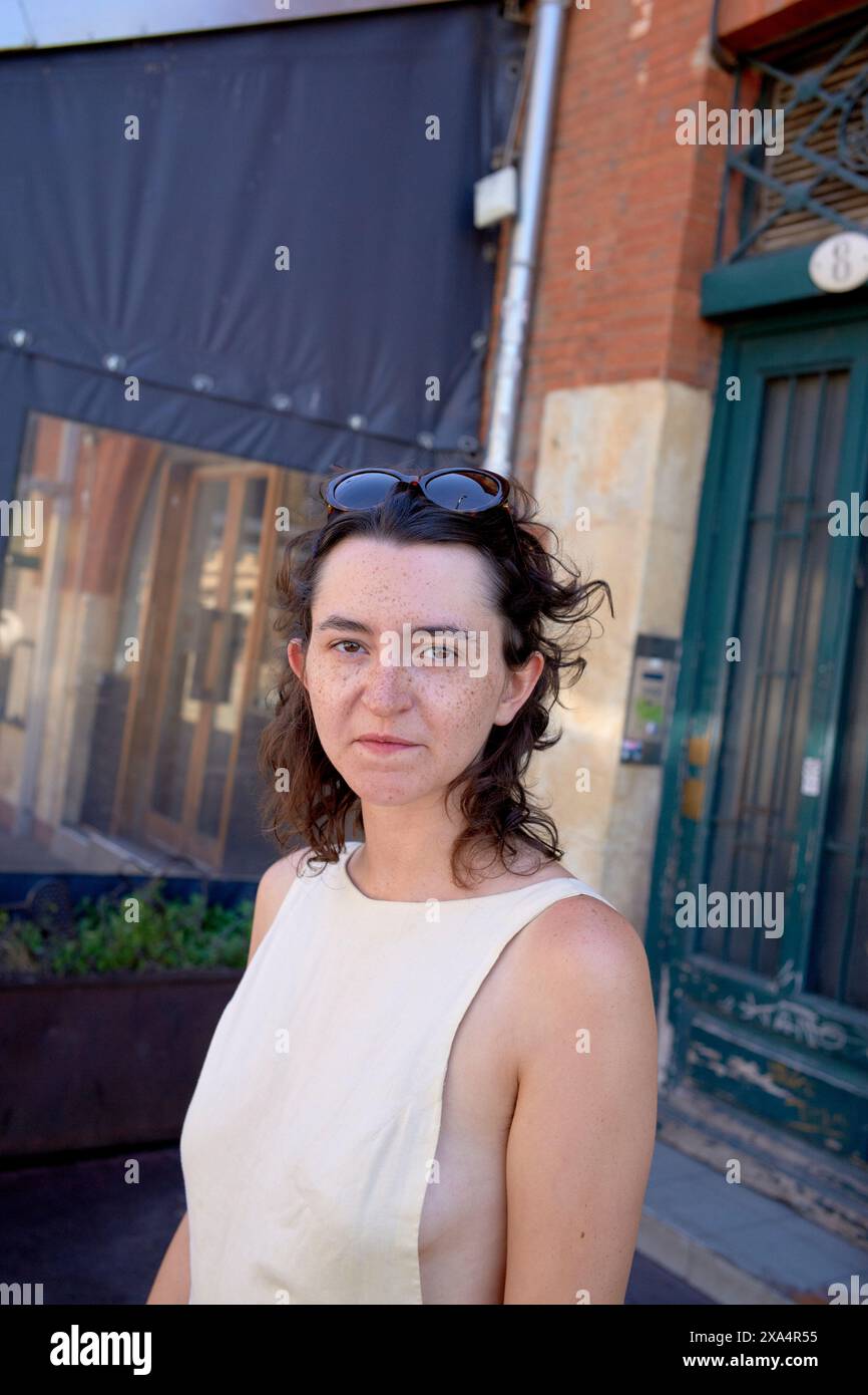 Eine Frau steht vor einem Backsteingebäude mit schwarzer Markise; sie trägt ein beigefarbenes ärmelloses Oberteil und eine Sonnenbrille auf dem Kopf. Stockfoto