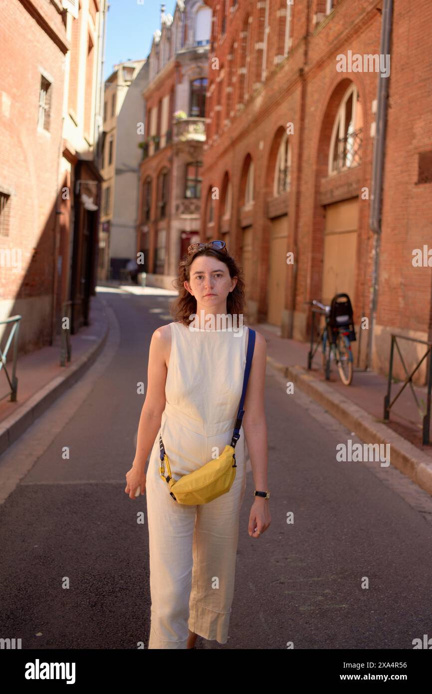 Eine Frau steht selbstbewusst in einer sonnenbeschienenen Gasse, trägt ein weißes ärmelloses Oberteil, eine cremefarbene Hose und eine gelbe Umhängetasche. Stockfoto