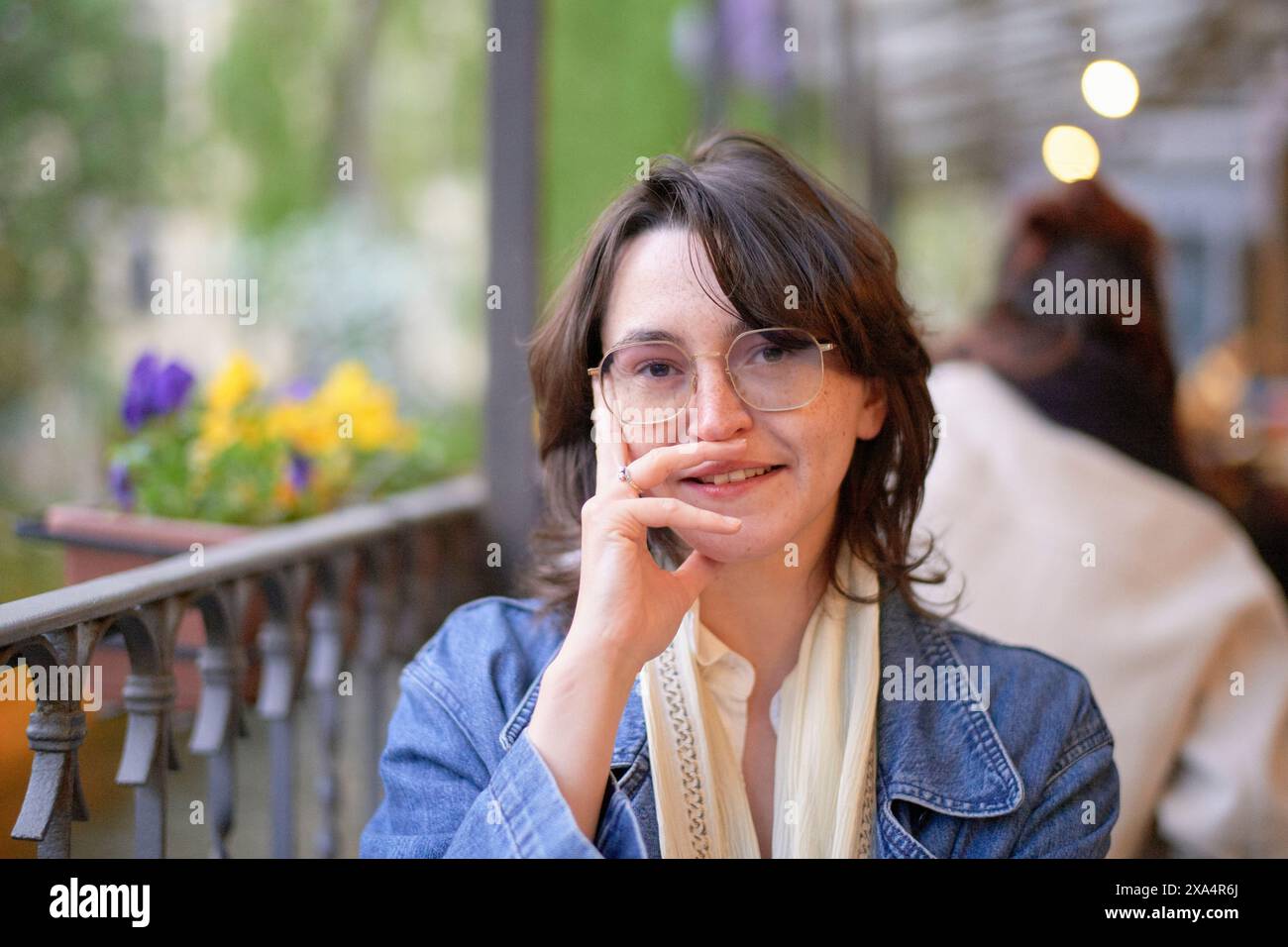 Eine lächelnde Frau mit Brille sitzt in einem Café im Freien und legt ihr Kinn auf ihre Hand. Stockfoto