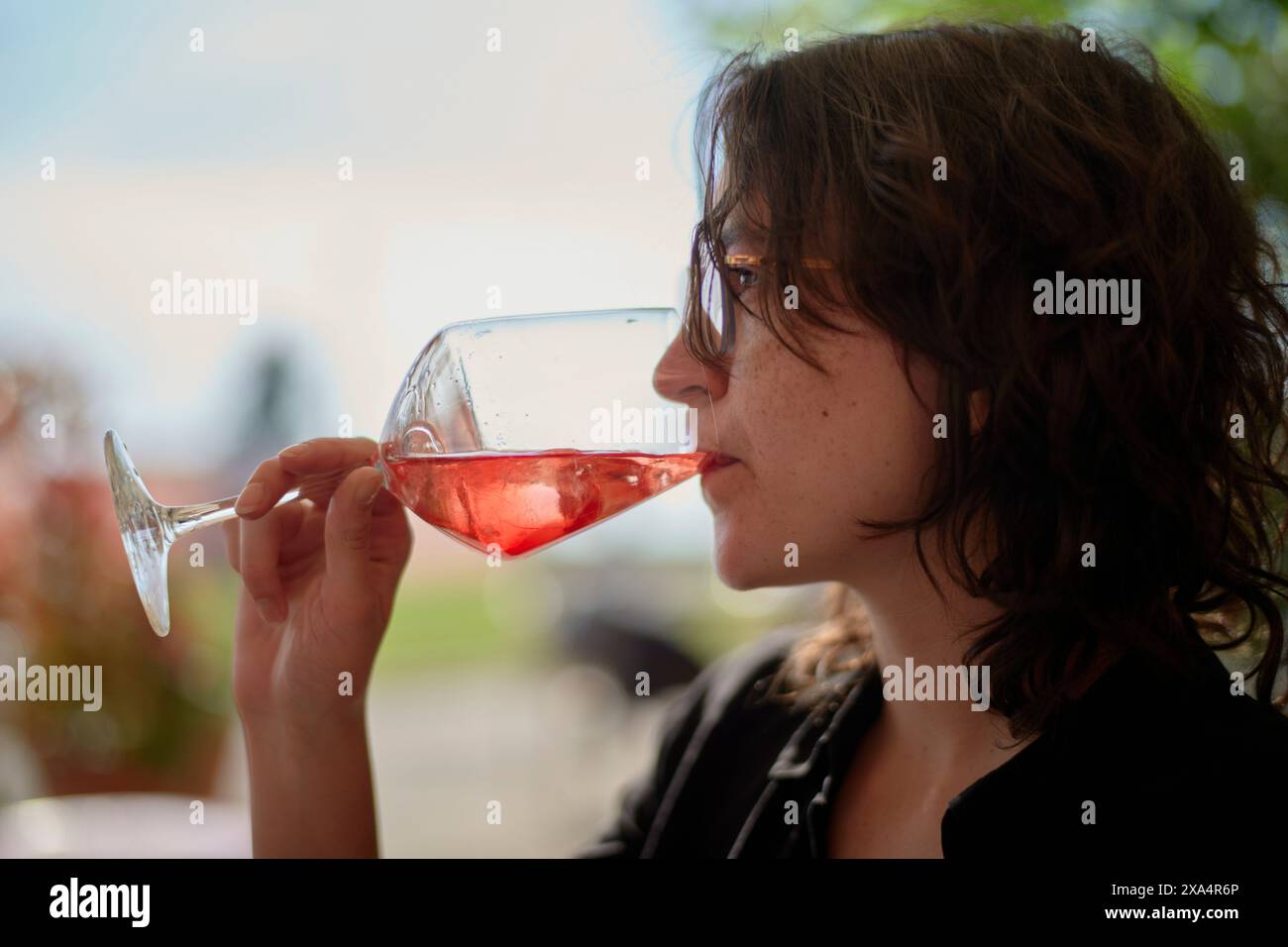 Eine junge Frau schlürft Rotwein aus einem Glas, mit weichem Fokus auf einem natürlichen Hintergrund. Stockfoto