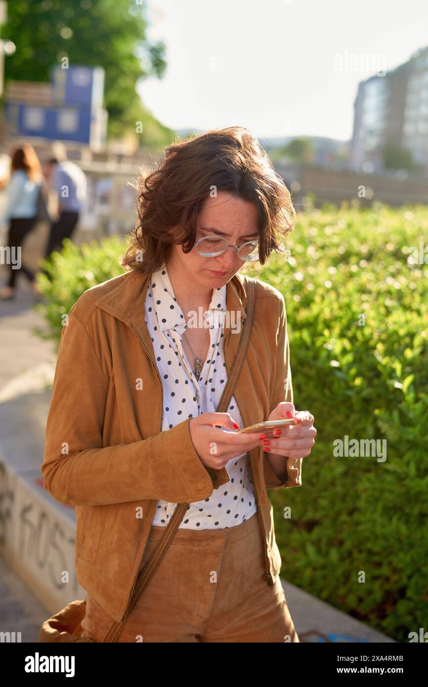 Frau in einer braunen Jacke, die an einem sonnigen Tag am Straßenrand auf ihrem Handy schreibt Stockfoto