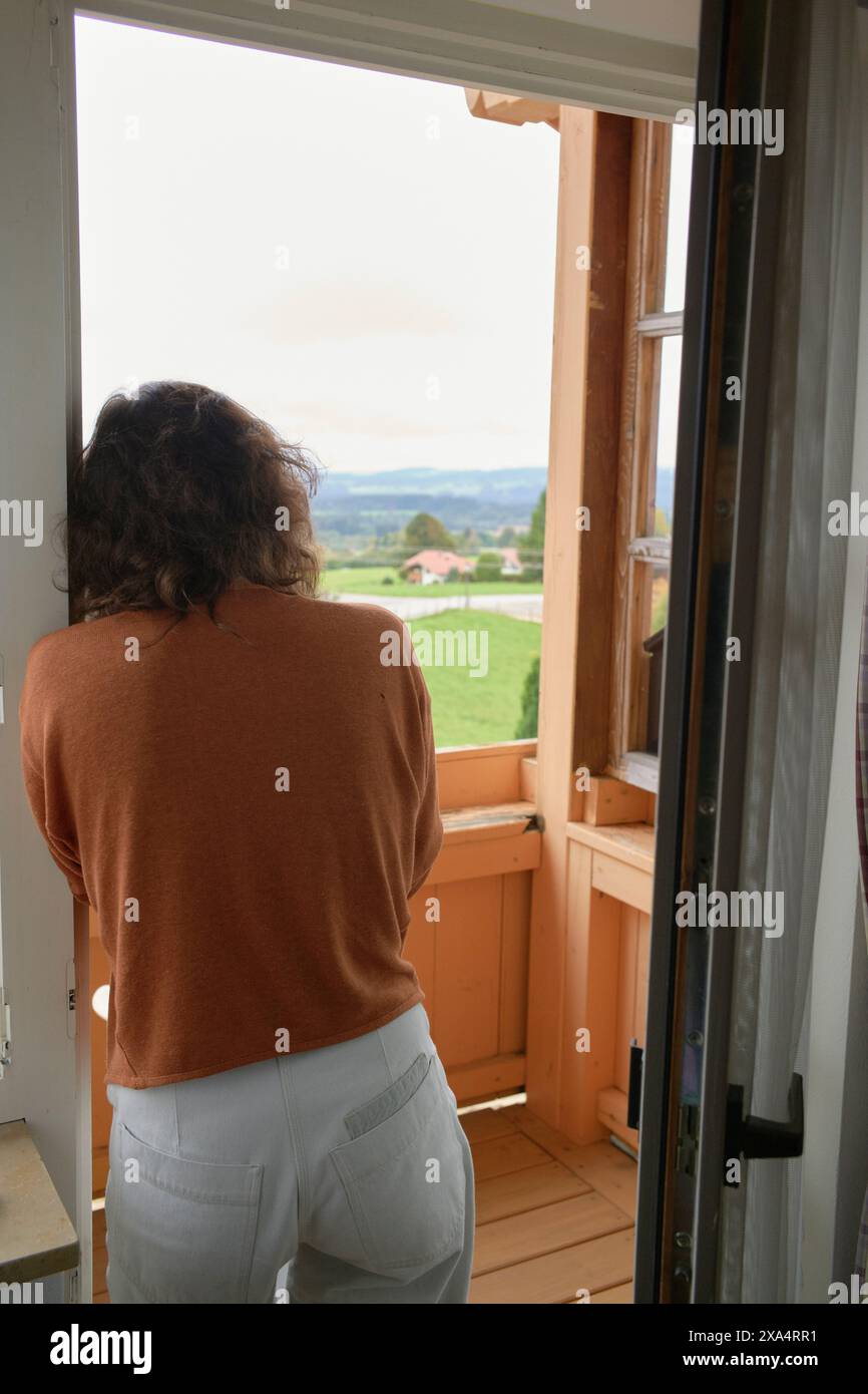 Frau, die aus einem offenen Fenster in eine landschaftlich reizvolle Landschaft blickt. Stockfoto